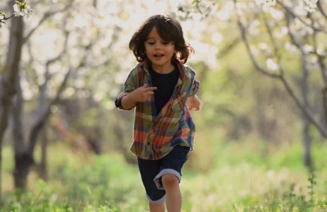 A child running outdoors along a grassy path surrounded by trees, wearing a plaid shirt and shorts, moving energetically through a bright natural setting.