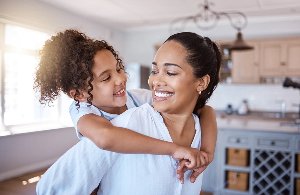 An adult carries a child on their back inside a bright kitchen, with the child’s arms wrapped around the adult’s shoulders.