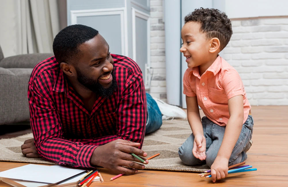 An adult and a young child are on the floor together with colored pencils, engaging in an art activity in a living room setting.