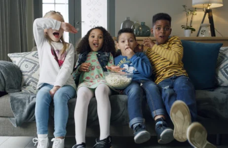 A group of four children sit together on a couch sharing a large bowl of popcorn in a living room.