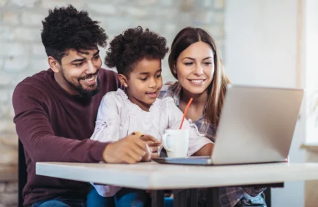 Three individuals sit closely together at a table while looking at a laptop screen. A cup with a straw and a notebook are on the table in front of them.