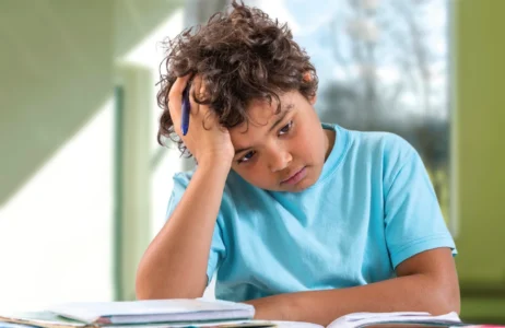 An individual wearing a light blue shirt sits at a table with an open notebook, resting their head on one hand while holding a pen. Books and papers are spread out on the table, and a bright window is visible in the background.