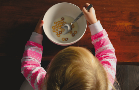 A child wearing pink polka‑dot pajamas sits at a table holding a bowl of cereal with milk. A spoon rests inside the bowl, which contains several round cereal pieces.