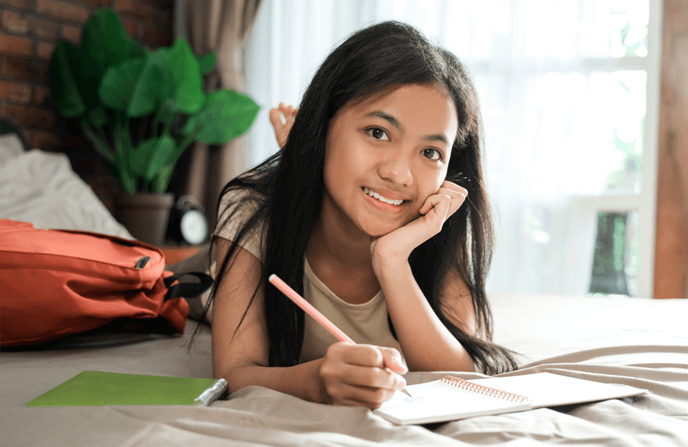 Teen girl smiling while journaling on her bed with a backpack nearby.