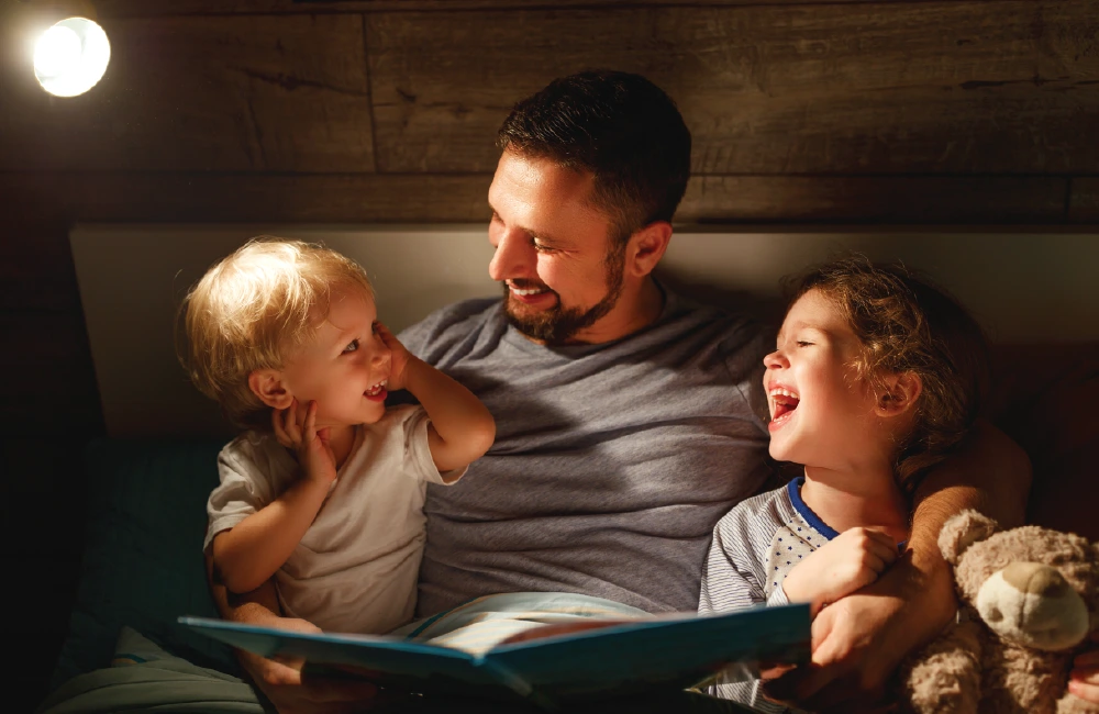 An adult sits in bed reading a book to two young children who are seated close on either side. A small light on the wall illuminates the cozy nighttime setting, and a stuffed animal is visible near one of the children.