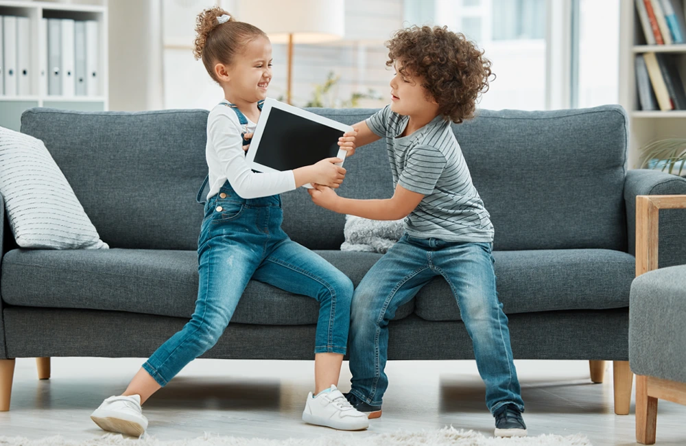Two individuals each carry a child on their back while walking outdoors in a park-like area with trees displaying golden leaves. The group is positioned on a pathway with soft natural lighting in the background.