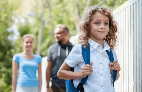 A child wearing a backpack walks outdoors along a sidewalk near a white fence, with two additional individuals walking behind. Trees and greenery appear in the background.