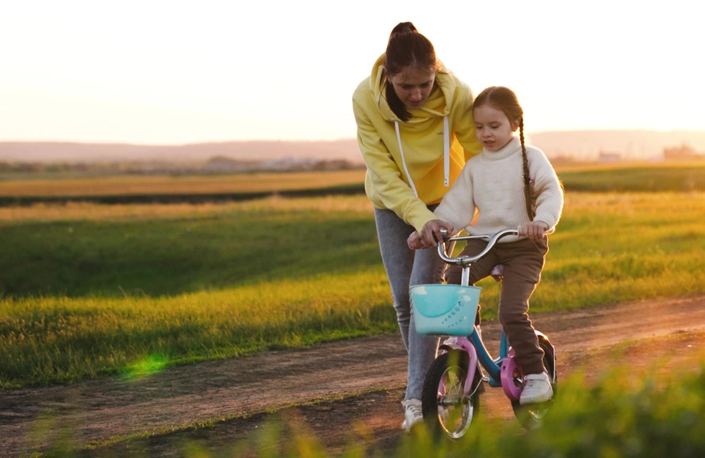 An adult walks beside a child who is learning to ride a small bicycle along an outdoor path at sunset.