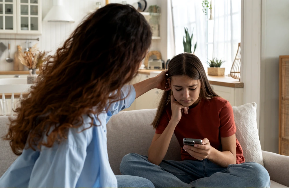An adult sits beside another person on a couch, gently placing a hand on the person’s head while the person holds a smartphone.