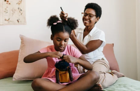 Adult gently parts and styles a child’s hair on a bed, while the child sits cross‑legged holding a small backpack.