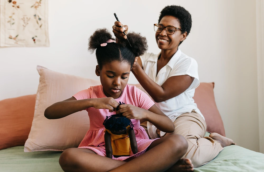 Adult gently parts and styles a child’s hair on a bed, while the child sits cross‑legged holding a small backpack.