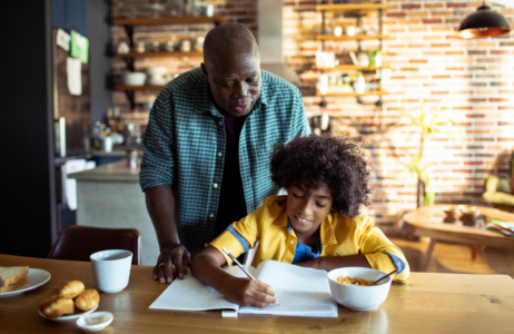 Adult standing beside a child at a kitchen table while the child writes in an open notebook.
