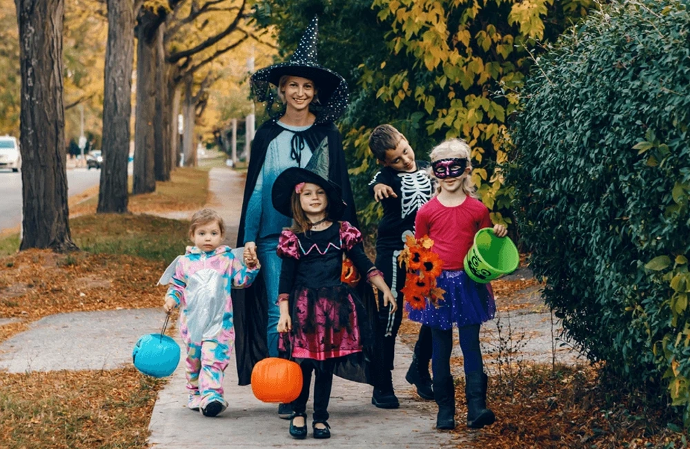 A group of children dressed in colorful Halloween costumes walk together along a tree-lined sidewalk, each carrying festive buckets for trick-or-treating.