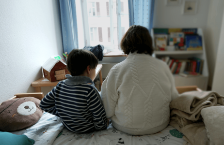 Two individuals sit side by side on a bed in a softly lit bedroom, facing a window. A bookshelf filled with books, a small table, and a round pillow are visible in the room.