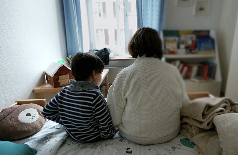 Two individuals sit side by side on a bed in a softly lit bedroom, facing a window. A bookshelf filled with books, a small table, and a round pillow are visible in the room.