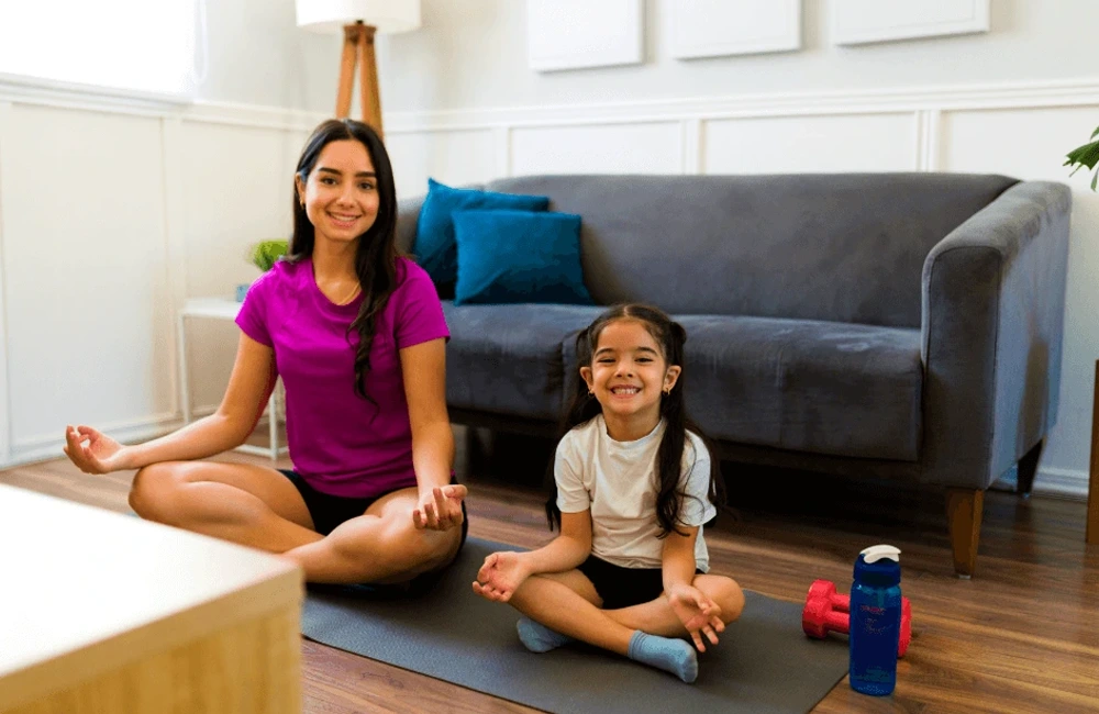 Two individuals sit on a yoga mat in a living room, practicing a seated meditation pose with legs crossed. A couch, pillows, dumbbells, and a water bottle are visible in the background.
