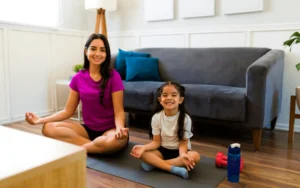 Two individuals sit on a yoga mat in a living room, practicing a seated meditation pose with legs crossed. A couch, pillows, dumbbells, and a water bottle are visible in the background.