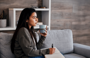 Thoughtful woman holding a mug and writing in a notebook while sitting on a gray couch.