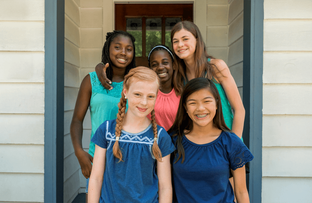A diverse group of five smiling girls standing together on the front porch of a house, looking happy and connected.