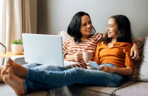 Parent and teenage daughter smiling together on a couch with a laptop and coffee, sharing a relaxed moment of connection and conversation.
