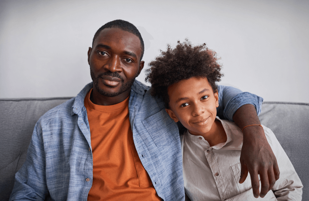 A father lovingly wraps his arm around his smiling son as they sit closely together on a couch, symbolizing connection and trust.