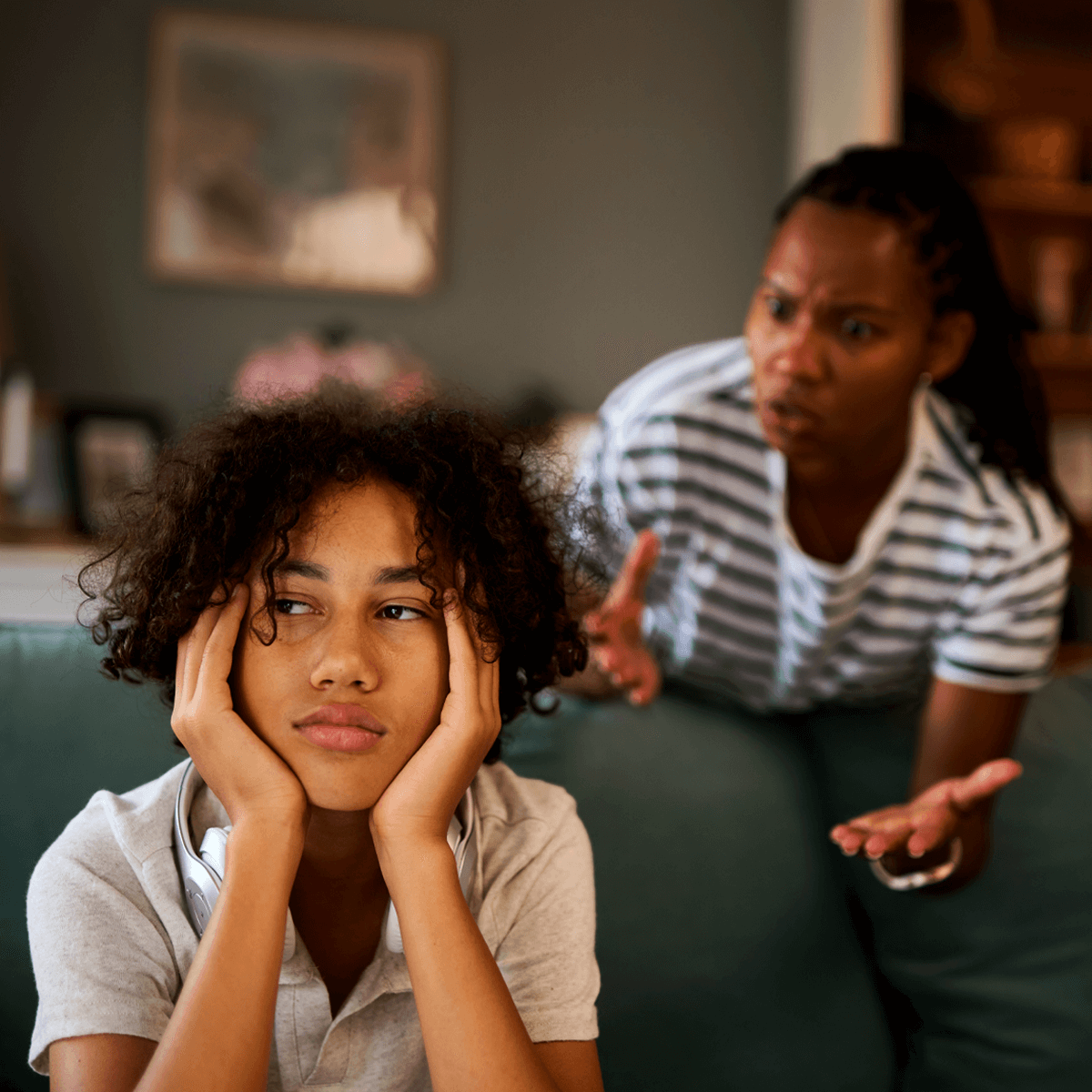 Teenager sits on a couch looking frustrated and withdrawn while an adult in the background appears to be speaking emotionally, illustrating the tension often experienced in trauma-affected relationships.