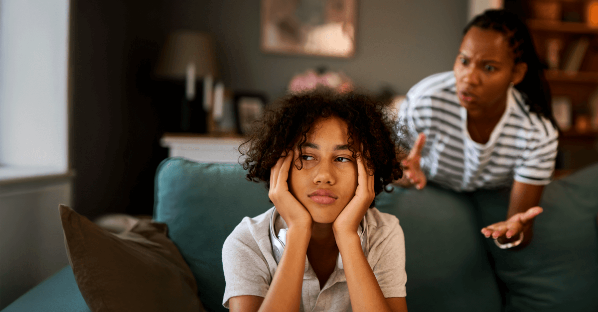 Teenager sits on a couch looking frustrated and withdrawn while an adult in the background appears to be speaking emotionally, illustrating the tension often experienced in trauma-affected relationships.
