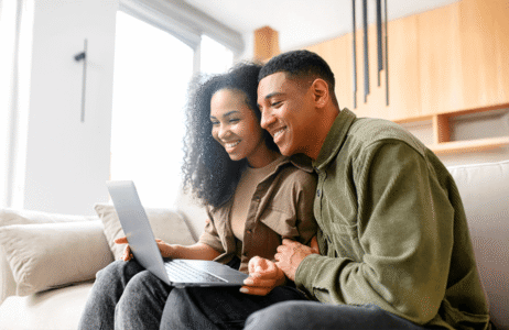 Smiling couple sitting together on a couch, looking at a laptop screen with interest and warmth, symbolizing connection and support in family relationships.