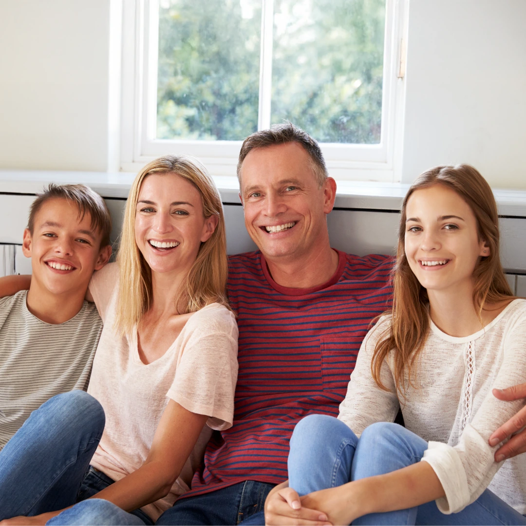 Smiling family of four sitting close together at home by a bright window—parents with two teens—warm, supportive environment.