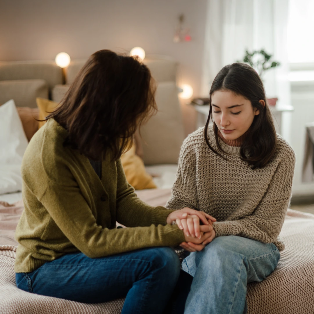 Two people sit together on a bed, holding hands in a moment of comfort and support, creating a warm, empathetic atmosphere.