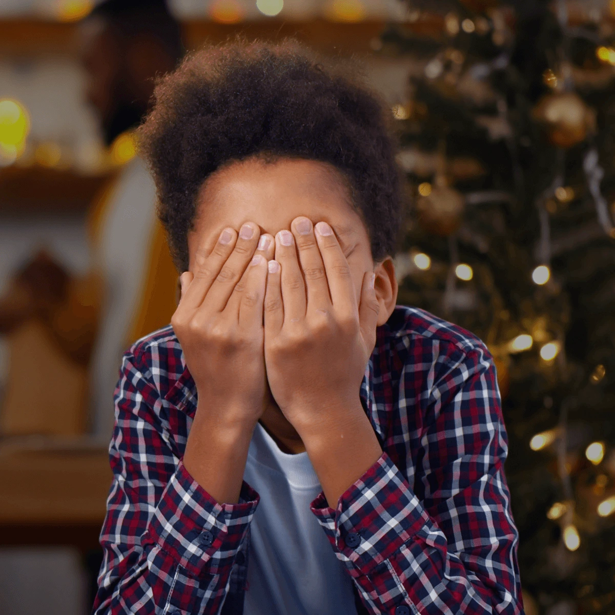 Child sitting in front of a decorated Christmas tree with hands covering face, wearing a plaid shirt, symbolizing holiday stress and overwhelm.