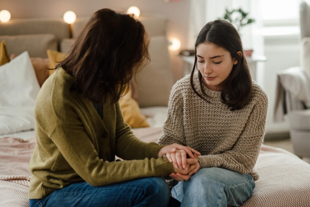A caregiver gently holds a teen’s hands during an emotional moment, modeling trauma-informed connection and support inside a cozy home environment.
