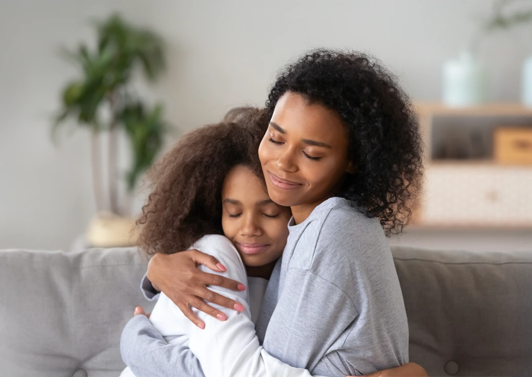 Mother and daughter sharing a tender hug on the couch, eyes closed in peace and connection.