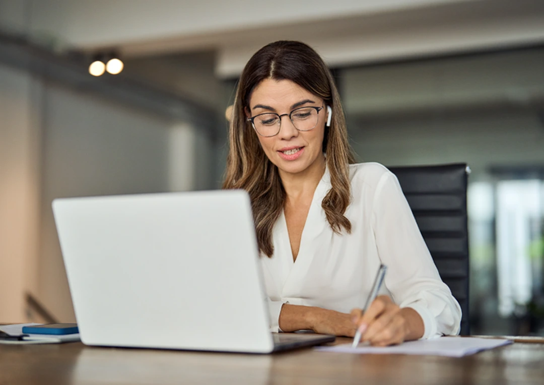 Professional woman working at a laptop and taking notes during an online meeting in a bright office.