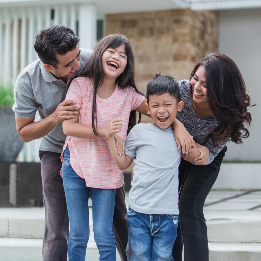 Smiling family of four linking arms and laughing together outside a home.