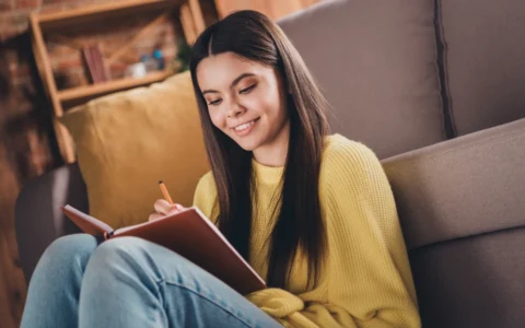 A teen sitting on a couch, smiling as she writes in a journal at home with guidance from ConnectionPlus resources.