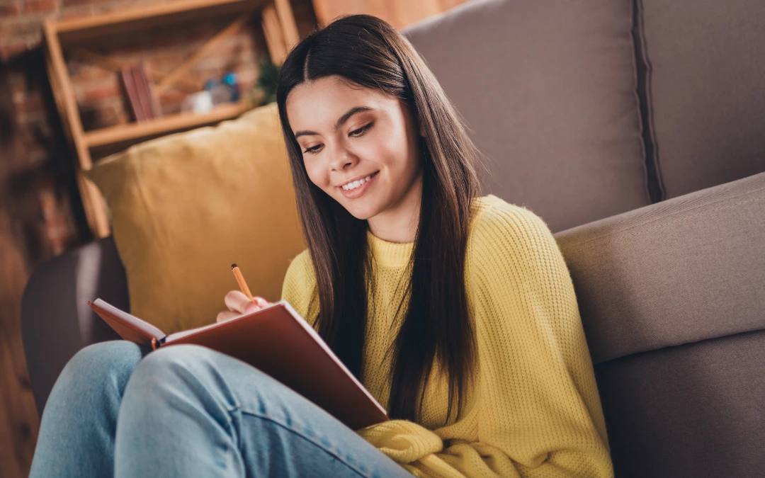A teen sitting on a couch, smiling as she writes in a journal at home with guidance from ConnectionPlus resources.