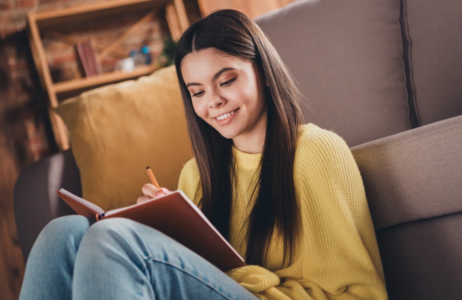 A teen sitting on a couch, smiling as she writes in a journal at home with guidance from ConnectionPlus resources.